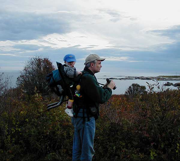 man with binoculars standing near ocean, with small child in carrier on his back
