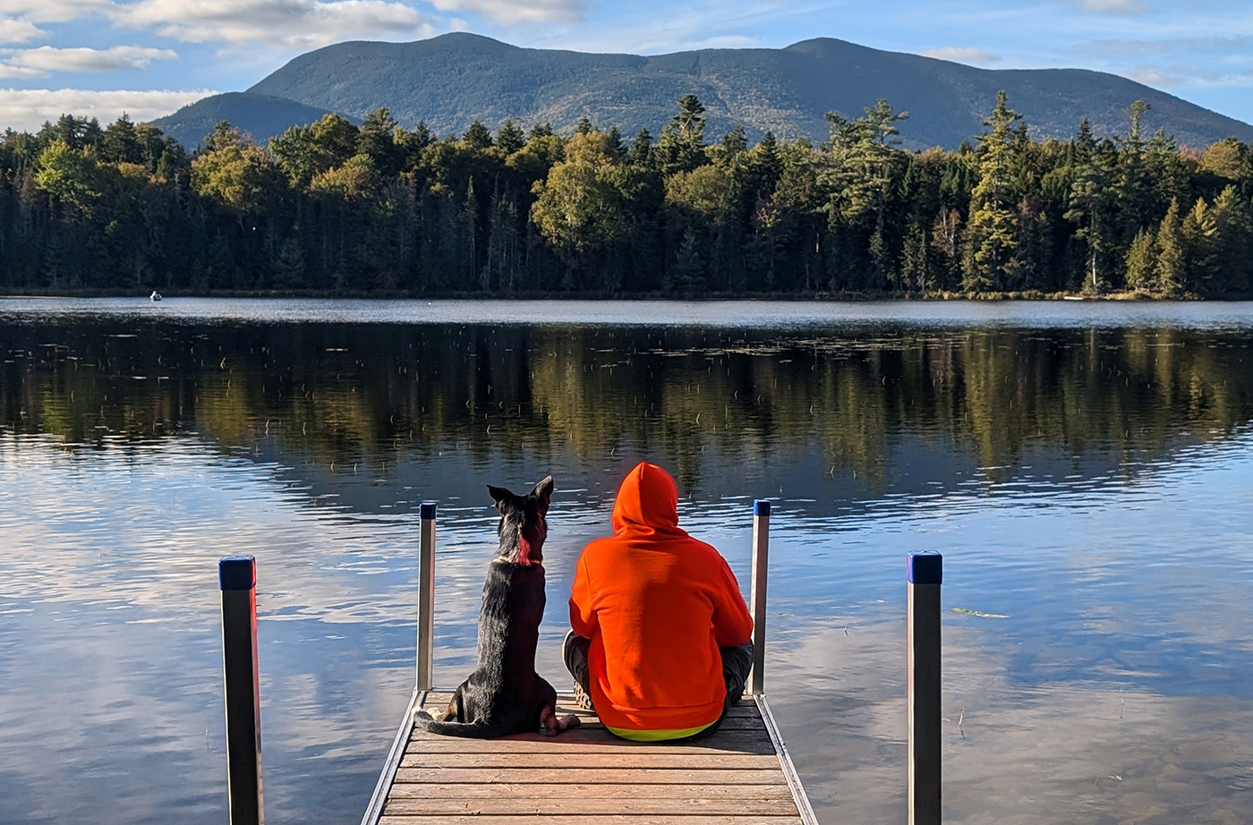 dog and person in hooded sweatshirt looking at mountain while sitting on wooden dock