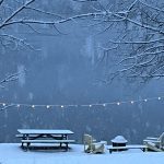 snowstorm over lake, with snow-covered picnic table, chairs, and firepit