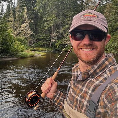 Nate Hathaway smiling wearing hat and sunglasses and holding a fly fishing rod